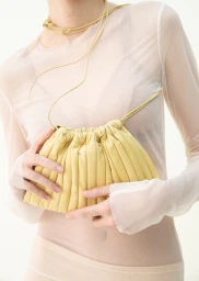 Model wearing a sheer off-white top holding a butter yellow pleated pouch bag against a white studio background.