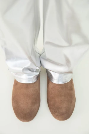 Top-down view of brown suede slip-on shoes paired with draped metallic silver trousers on a white studio floor.