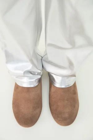 Top-down view of brown suede slip-on shoes paired with draped metallic silver trousers on a white studio floor.