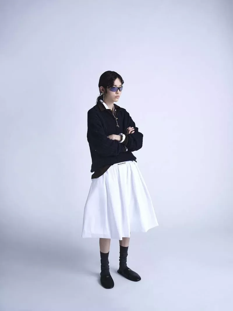 A person stands in a photo studio wearing a black long sleeve shirt, white skirt, socks, and shoes in a studio setting.
