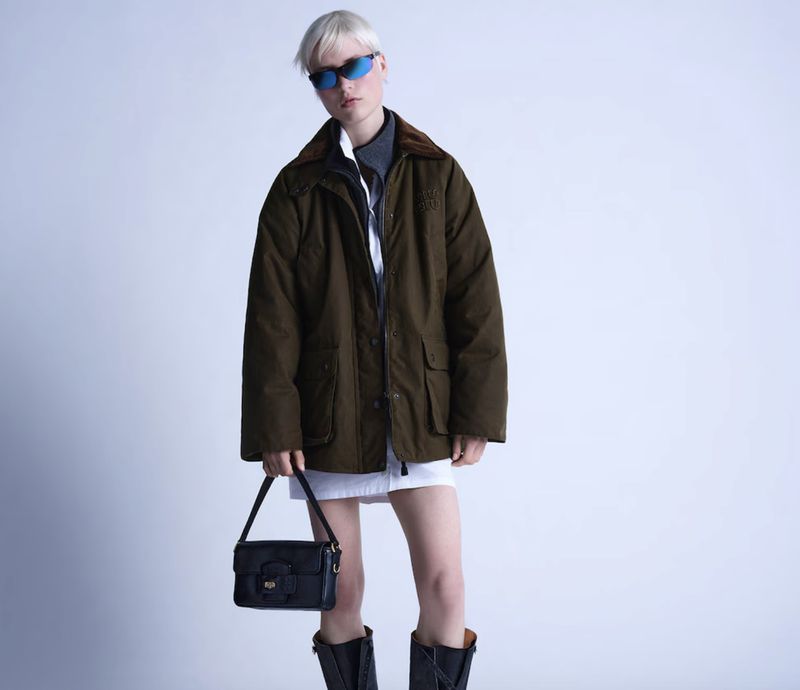 Fashionable woman in green jacket, white skirt, and black boots poses indoors with a purse, against a clean studio background.