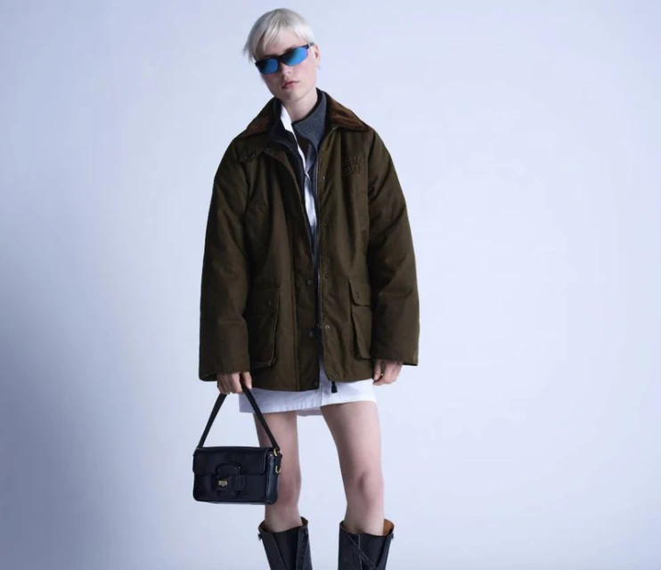 Fashionable woman in green jacket, white skirt, and black boots poses indoors with a purse, against a clean studio background.