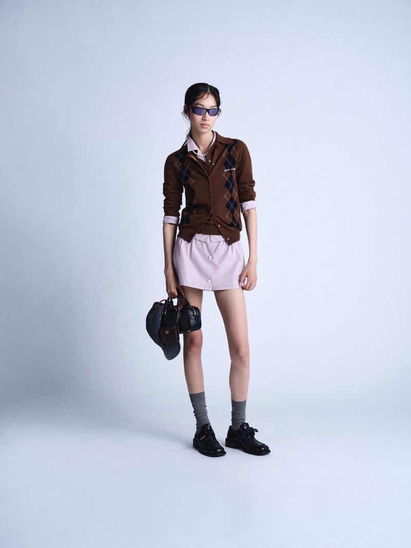 Full-length shot of a woman in a brown argyle cardigan, lilac skirt, socks, and black shoes, holding a black bag, against a grey background.