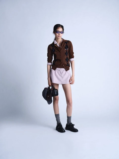 Full-length shot of a woman in a brown argyle cardigan, lilac skirt, socks, and black shoes, holding a black bag, against a grey background.