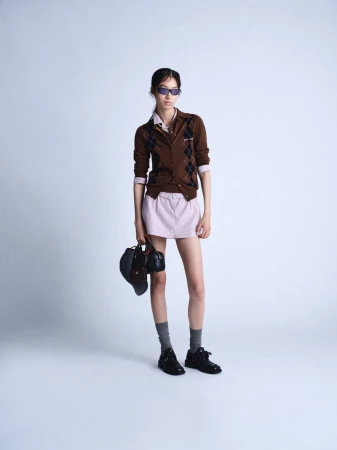Full-length shot of a woman in a brown argyle cardigan, lilac skirt, socks, and black shoes, holding a black bag, against a grey background.