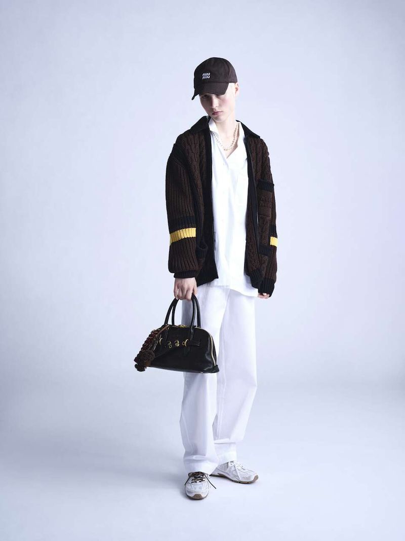 Full-shot of a young adult wearing a brown cardigan, white shirt and pants, a dark baseball cap, and holding a dark leather bag.