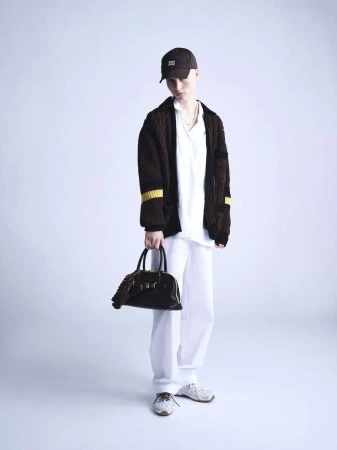 Full-shot of a young adult wearing a brown cardigan, white shirt and pants, a dark baseball cap, and holding a dark leather bag.