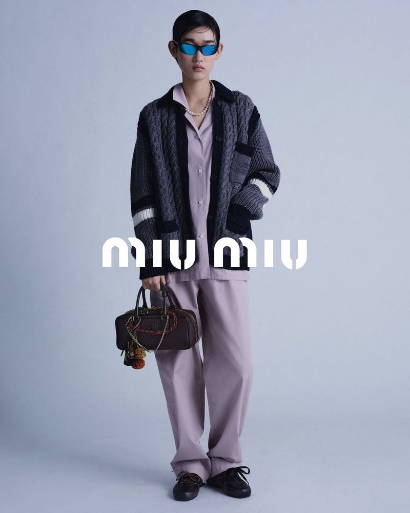 Full shot of a fashion model wearing a lilac outfit and dark cardigan, holding a brown handbag with the Miu Miu logo visible.