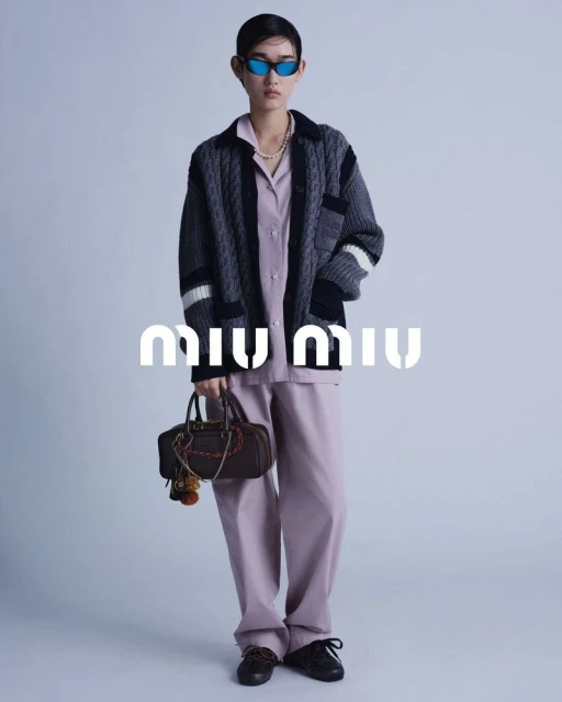 Full shot of a fashion model wearing a lilac outfit and dark cardigan, holding a brown handbag with the Miu Miu logo visible.