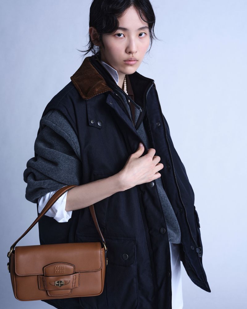 Model in dark jacket and white shirt holding a brown shoulder bag against a neutral studio backdrop