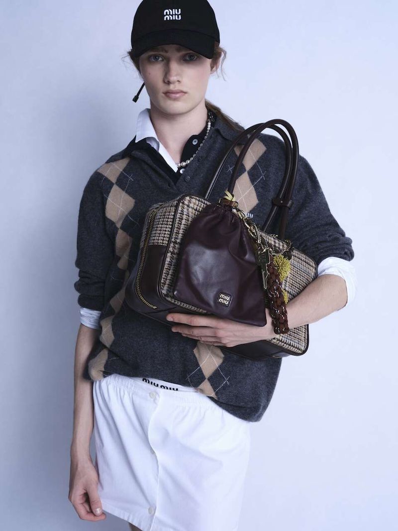 Fashion shot of a male model wearing Miu Miu cap and argyle sweater, holding a purse in a studio with a neutral backdrop.