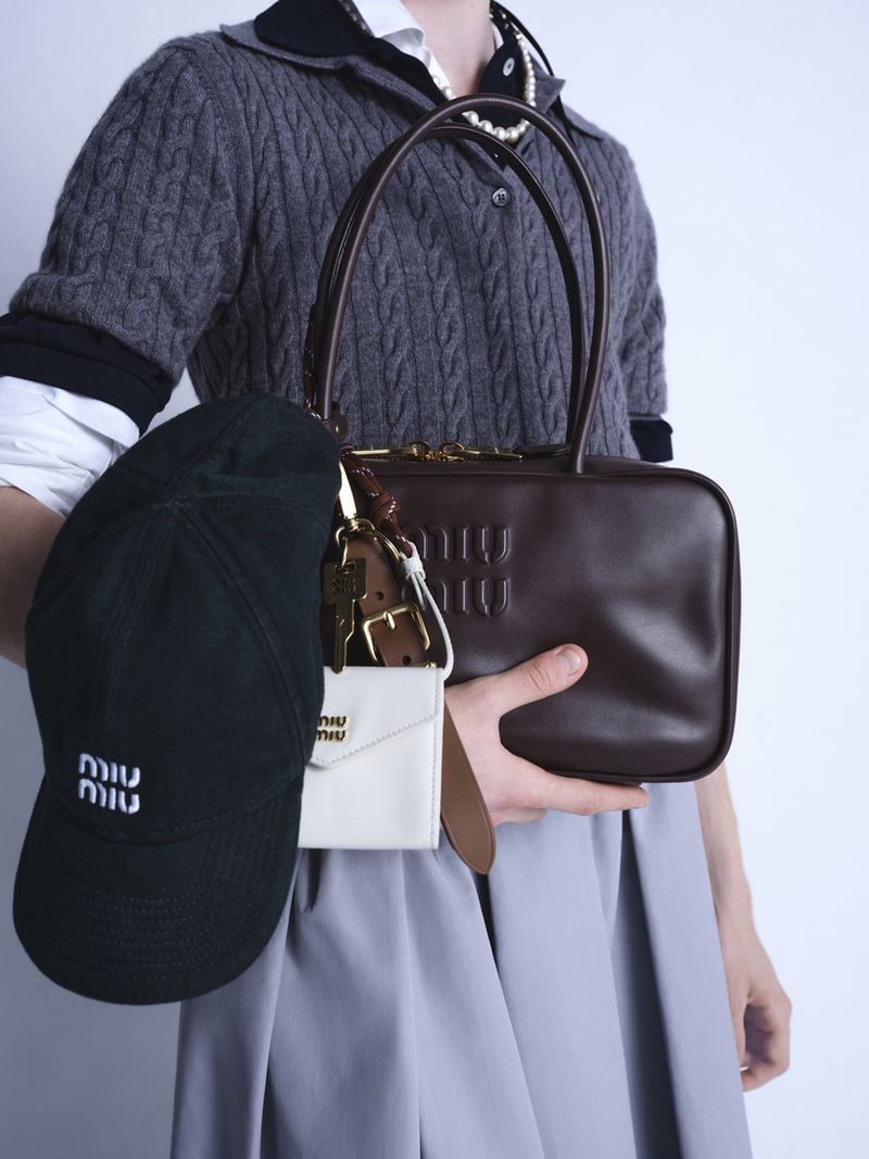 Person holding Miu Miu brown leather bag, black baseball cap, and white card holder against a pale gray studio backdrop.
