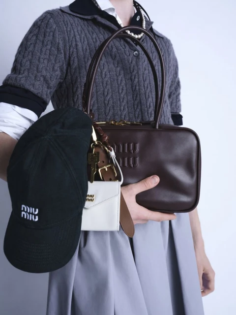 Person holding Miu Miu brown leather bag, black baseball cap, and white card holder against a pale gray studio backdrop.