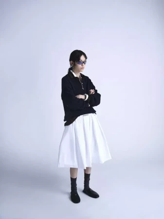 Model with sunglasses wearing a black polo shirt and white pleated skirt against a white studio background