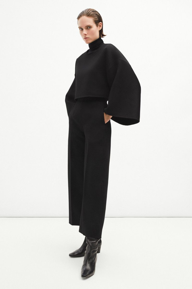 Fashion model posing in black turtleneck top, wide-leg pants, and black boots against a white background in a studio shot.