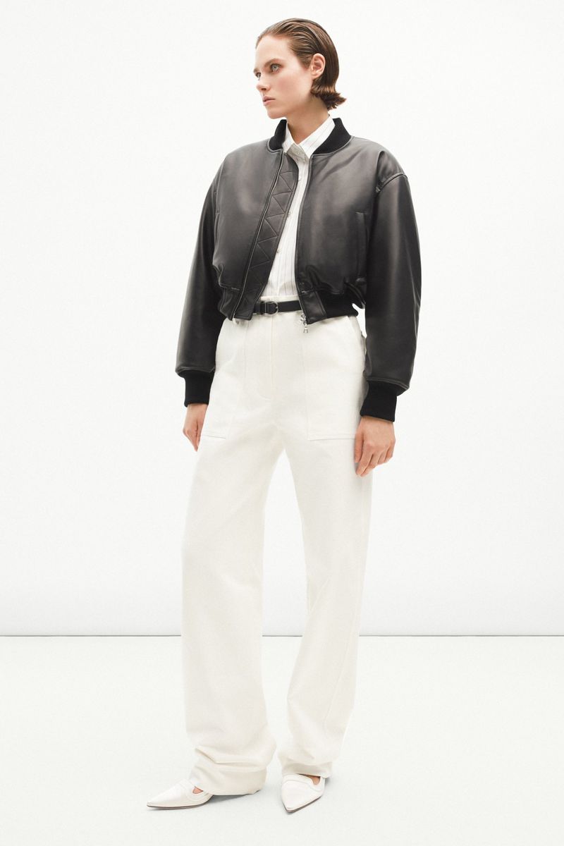 Fashionable woman in a cropped leather bomber jacket, white wide-leg pants, and white heels, posing in a minimalist studio setting.