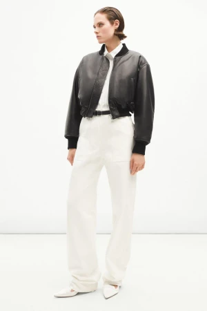 Fashionable woman in a cropped leather bomber jacket, white wide-leg pants, and white heels, posing in a minimalist studio setting.