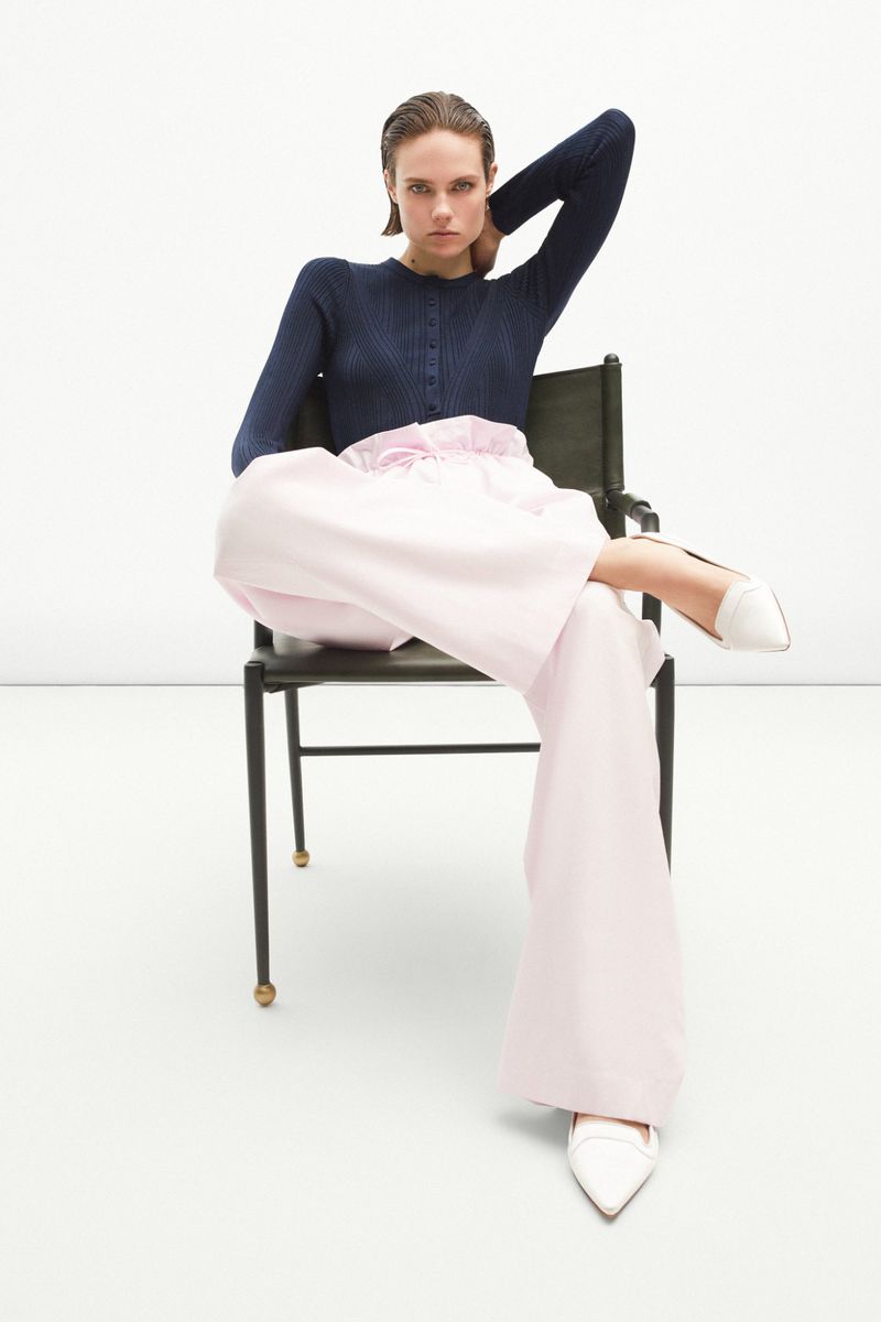 Adult woman poses on a chair wearing a navy button-down cardigan, wide-leg pink trousers, and white pointed-toe shoes in a studio.