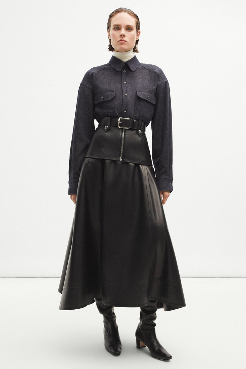 Fashion portrait of a woman in a navy blouse, black skirt with belt, and black boots, standing against a white backdrop.