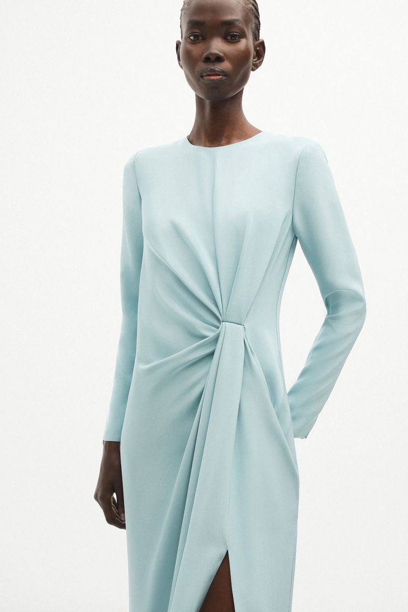 Elegant adult woman in a light blue, long-sleeve midi dress posing in a minimalist white studio setting for a fashion editorial shot.