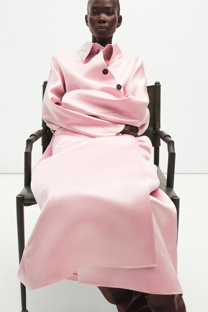 Studio shot of a Black female model in a pink coat, seated in a black wood chair with arms crossed against a plain, bright backdrop.