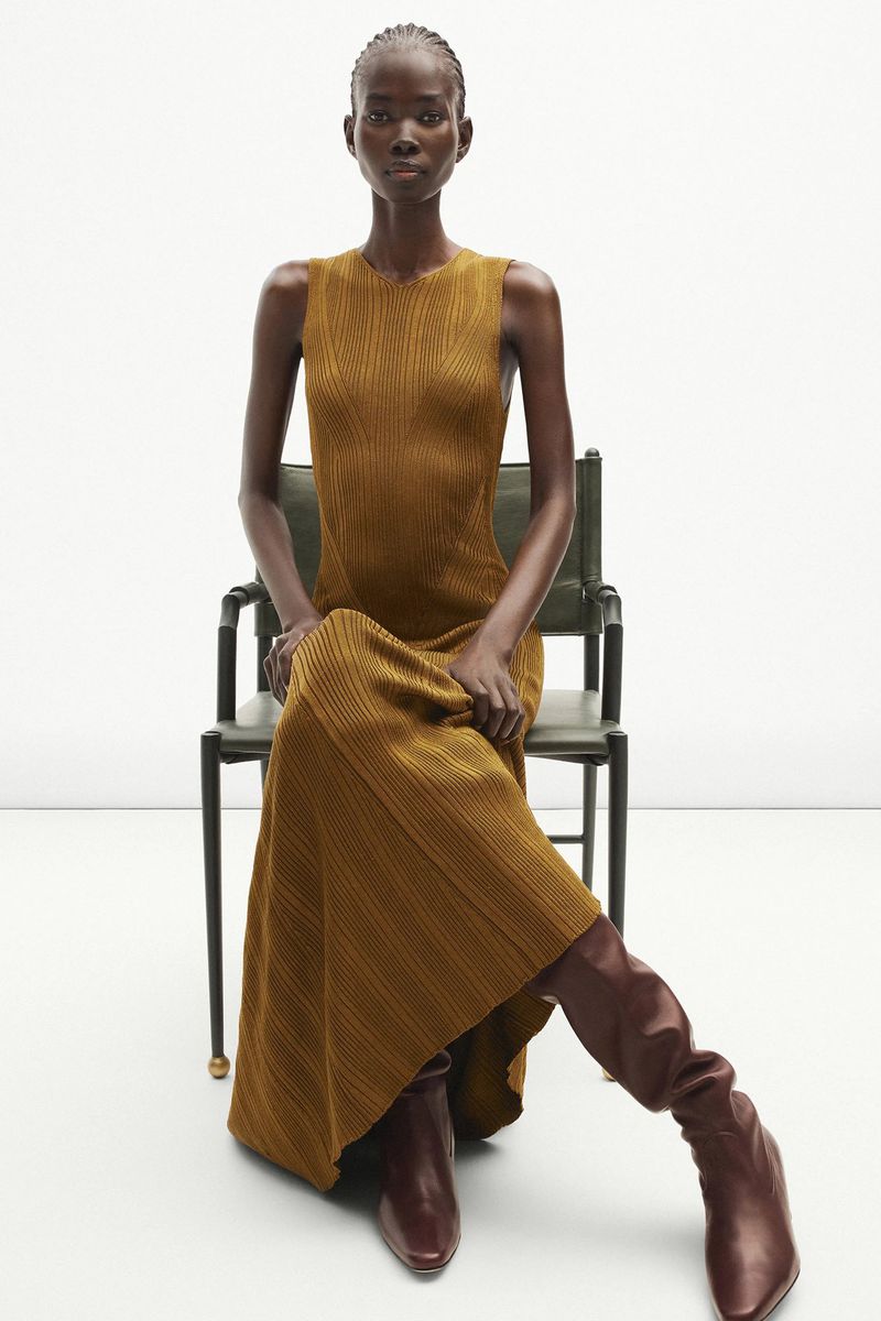 An African-American woman in a gold dress and leather boots poses seated on a chair against a white backdrop in a fashion setting.