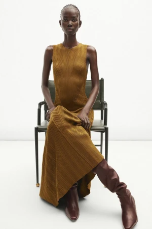 Model Mammina Aker seated in a sleeveless mustard knit maxi dress and tall leather boots for Adam Lippes Resort 2025.