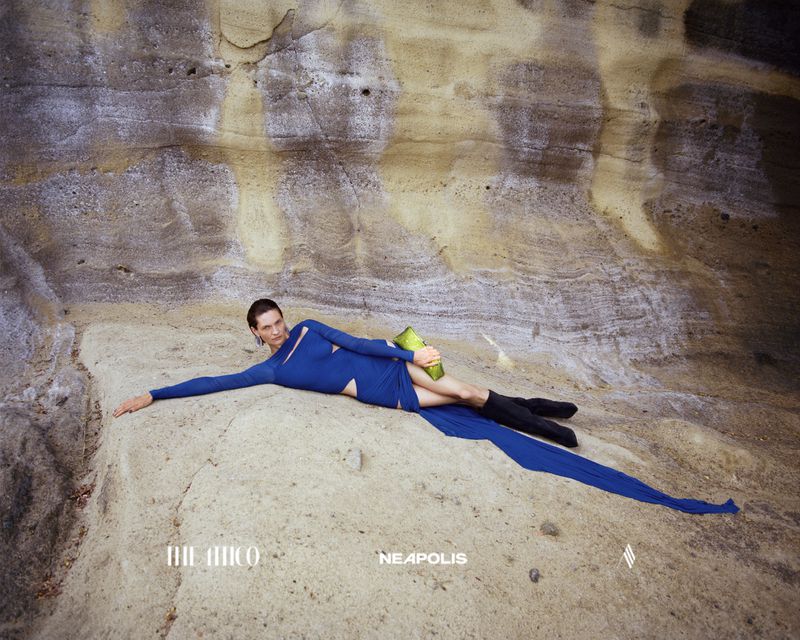 A stylish person in a blue cut-out dress poses on sandstone in a cave, holding a neon green object. Fashion editorial photography.