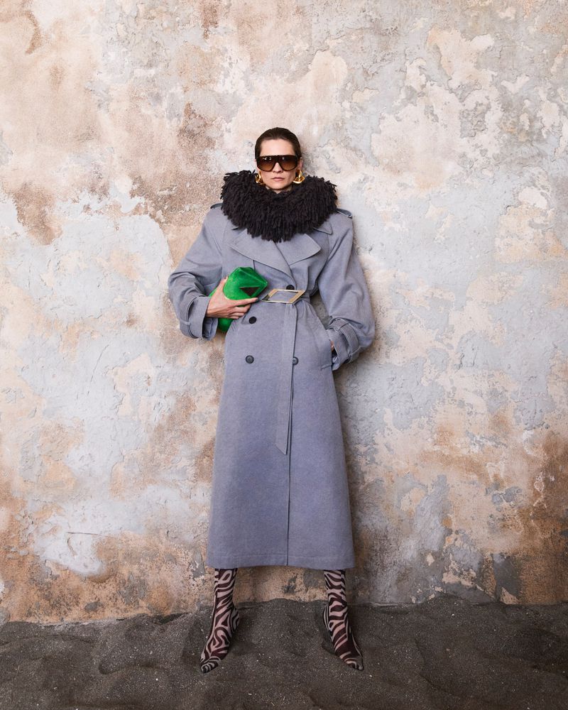 A woman in a grey coat, black collar, and patterned boots stands indoors holding a green clutch, posing against a textured wall.