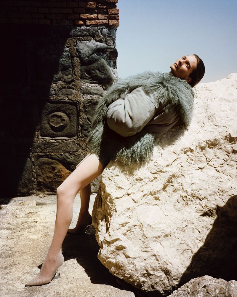 Fashion shot of a woman wearing a furry coat, posing next to a rock wall and rock, under a bright sky, outdoors.