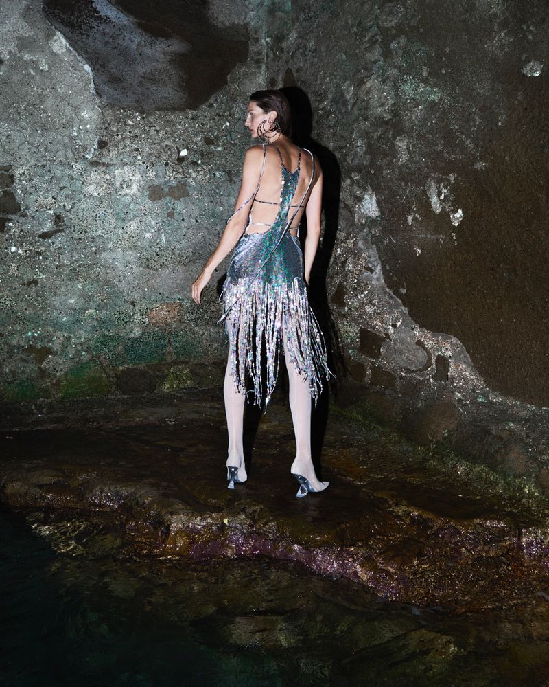 Back view of a slender adult female in a shimmering silver dress with fringe, posing in a dark, textured, cavern-like space with water reflections.