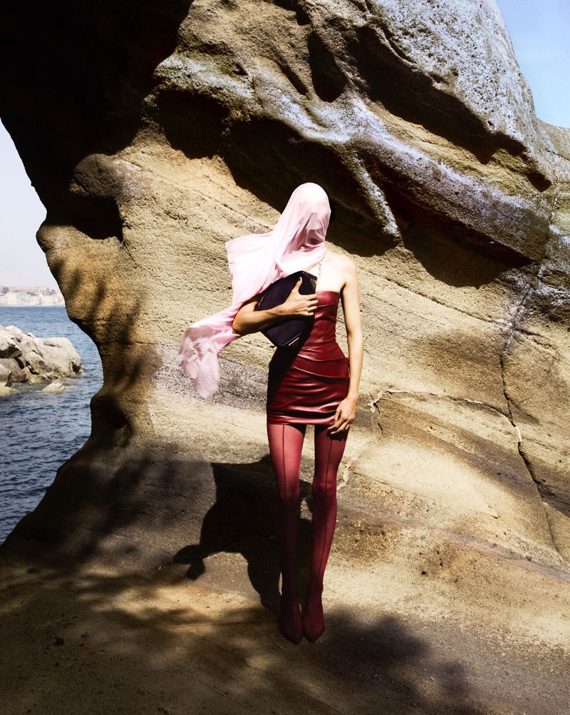 Fashion shot of a woman in a red dress and pink headscarf standing near a rock formation by the sea on a sunny day.