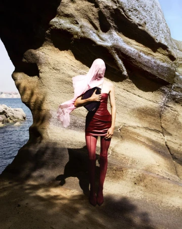 Fashion shot of a woman in a red dress and pink headscarf standing near a rock formation by the sea on a sunny day.