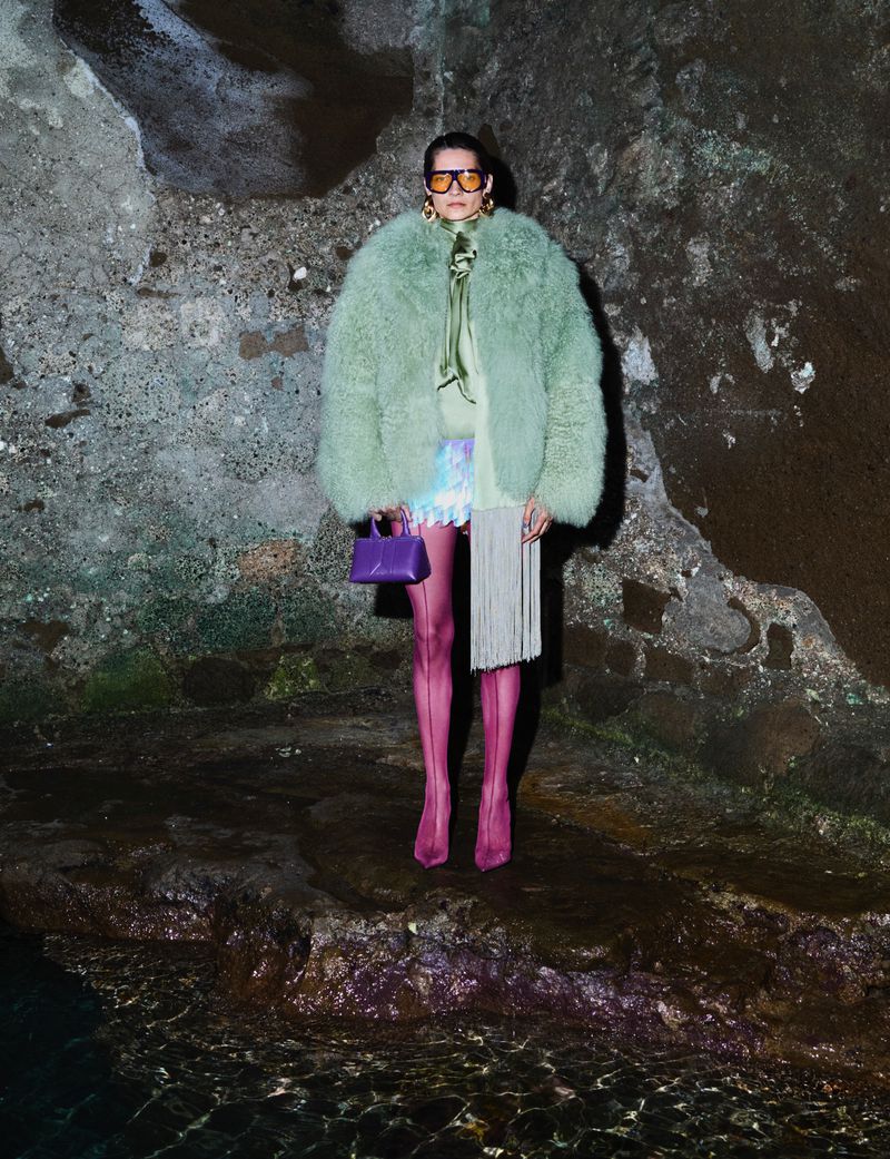 Fashionable adult female posing in a fur coat with handbag, sunglasses, and pink stockings in front of a textured wall.