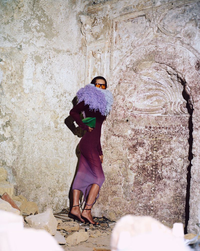 Woman in ombre dress and purple collar stands in stone ruins with green bag, strappy sandals, and amber sunglasses in a fashion shot.