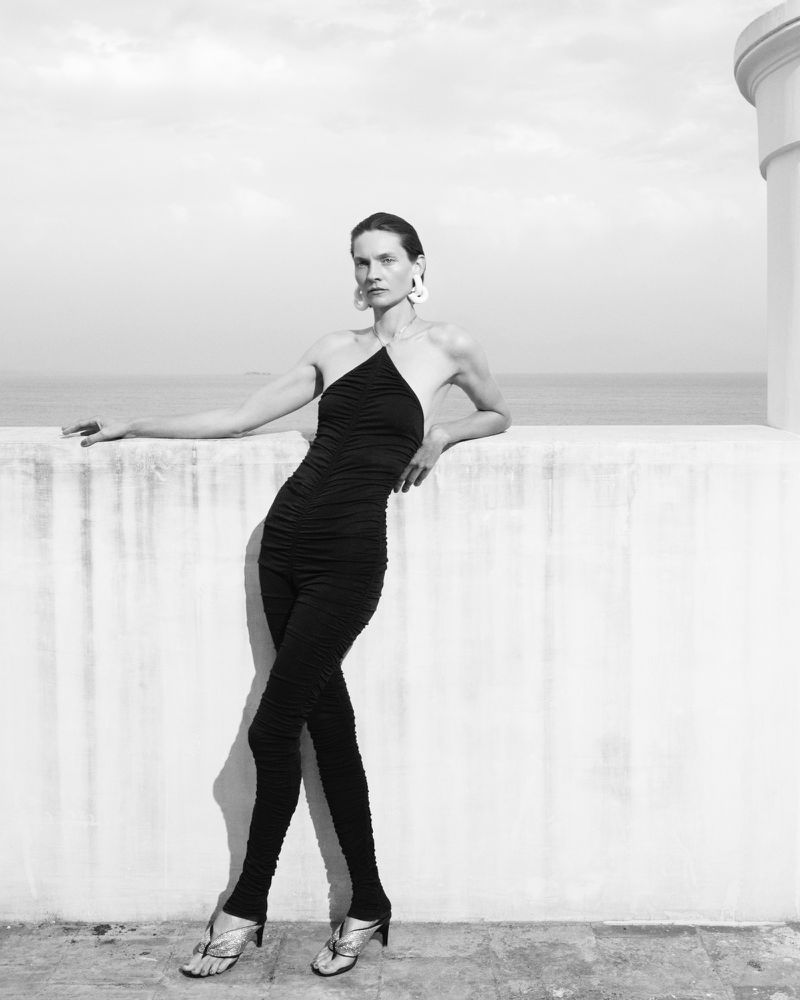 Black-and-white shot of an adult female fashion model wearing a one-shoulder jumpsuit and posing outdoors against the backdrop of a sea view.