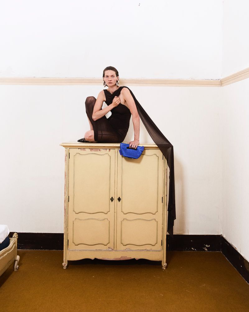 Adult female posing on a cabinet in a photo studio, wearing a dark top and sheer tights with black fabric draped over her arm.