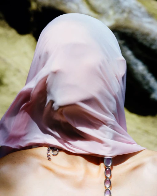 Surreal image featuring an adult female draped with a sheer pink cloth, wearing a jeweled necklace outdoors against a soft beige backdrop.