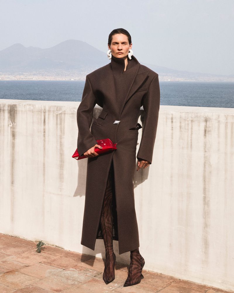 Fashion portrait of a stylish adult woman in a long brown coat holding a red clutch, posing outdoors with an ocean and mountain background.
