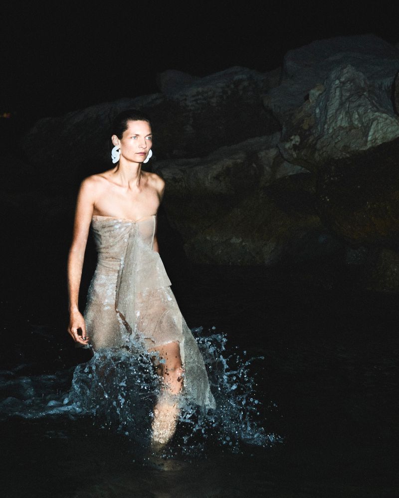 Woman in flowing dress walking through water, illuminated against a dark background with rocks and splashing water.