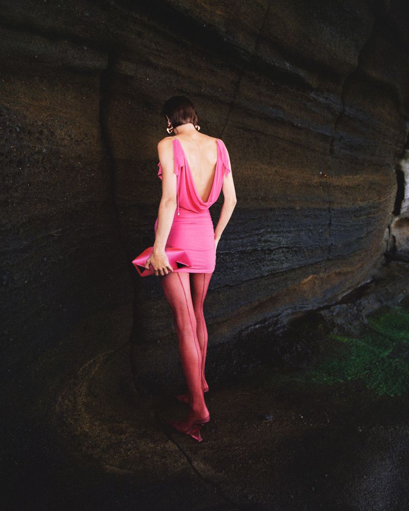 Back view of a female fashion model in a bright pink dress and pink tights, holding a matching purse and posing in an outdoor location.