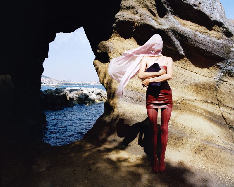Woman in red skirt and tights stands near rock formation, face obscured by pink veil, with ocean and city in background on a sunny day.