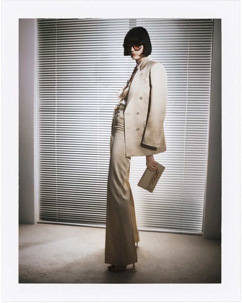 Elegant woman in a beige suit and sunglasses poses indoors in front of window blinds, holding a clutch in a full-length fashion shot.