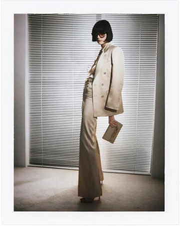 Elegant woman in a beige suit and sunglasses poses indoors in front of window blinds, holding a clutch in a full-length fashion shot.