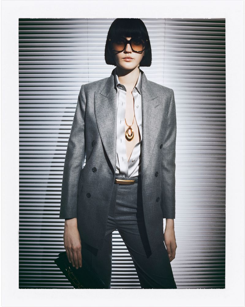 A woman in a grey blazer and matching trousers poses against a backdrop of horizontal blinds, wearing sunglasses and a statement necklace.