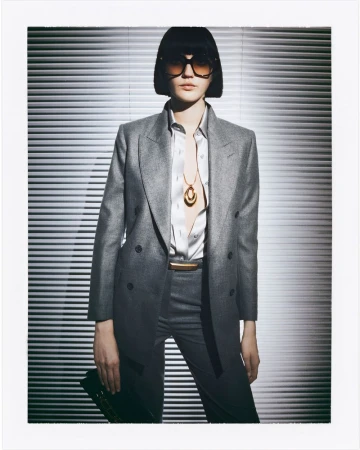 A woman in a grey blazer and matching trousers poses against a backdrop of horizontal blinds, wearing sunglasses and a statement necklace.