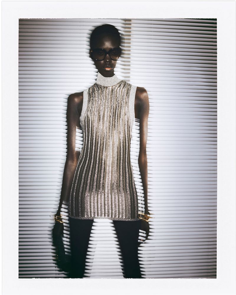 A slender Black woman poses in a sparkly gold top, black leggings, gold bracelets, and dark glasses in front of white blinds.