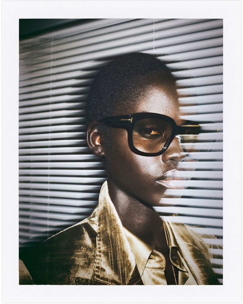 Close-up of a model with dark skin wearing black-framed glasses and a shiny gold jacket in front of horizontal window blinds.