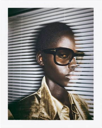 Close-up of a model with dark skin wearing black-framed glasses and a shiny gold jacket in front of horizontal window blinds.