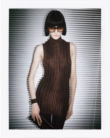 A young adult female poses indoors in a sleeveless, ribbed, metallic brown top, black pants, and dark sunglasses, in front of silver blinds.